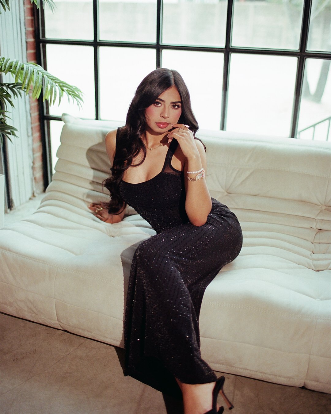 A woman with long dark hair in a black, sparkly dress sitting stylishly on a cream-colored sofa in a bright room with large windows, a brick wall, and a potted plant.