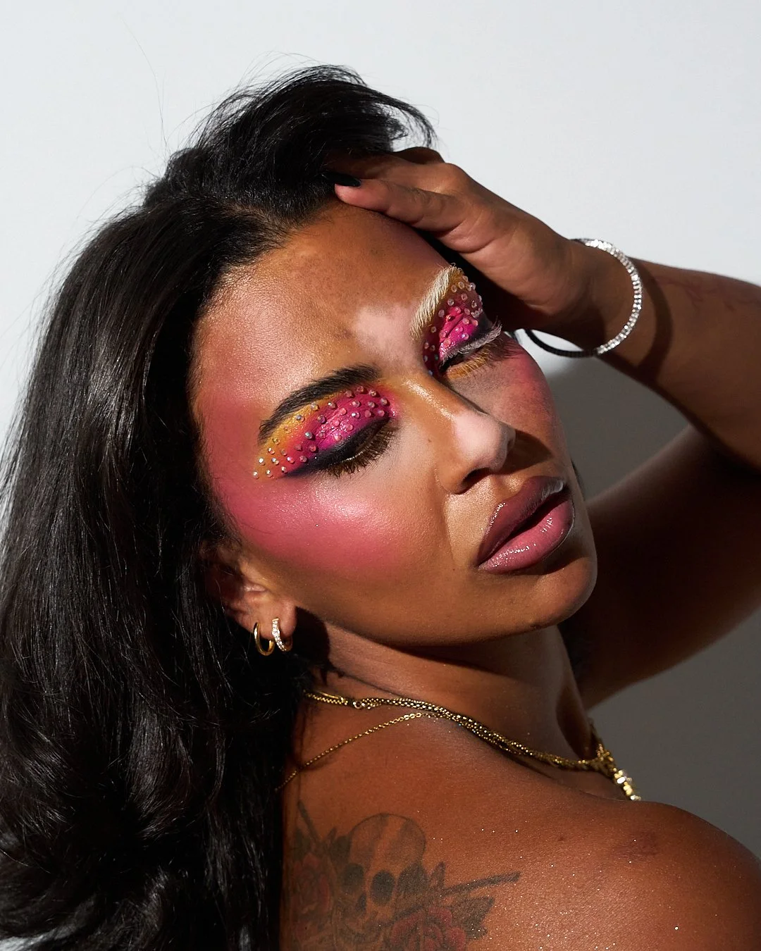 Portrait of a woman with elaborate eye makeup featuring pink and yellow eyeshadow, decorated with rhinestones, and pink blush, wearing gold jewelry and tattoos on her shoulder.
