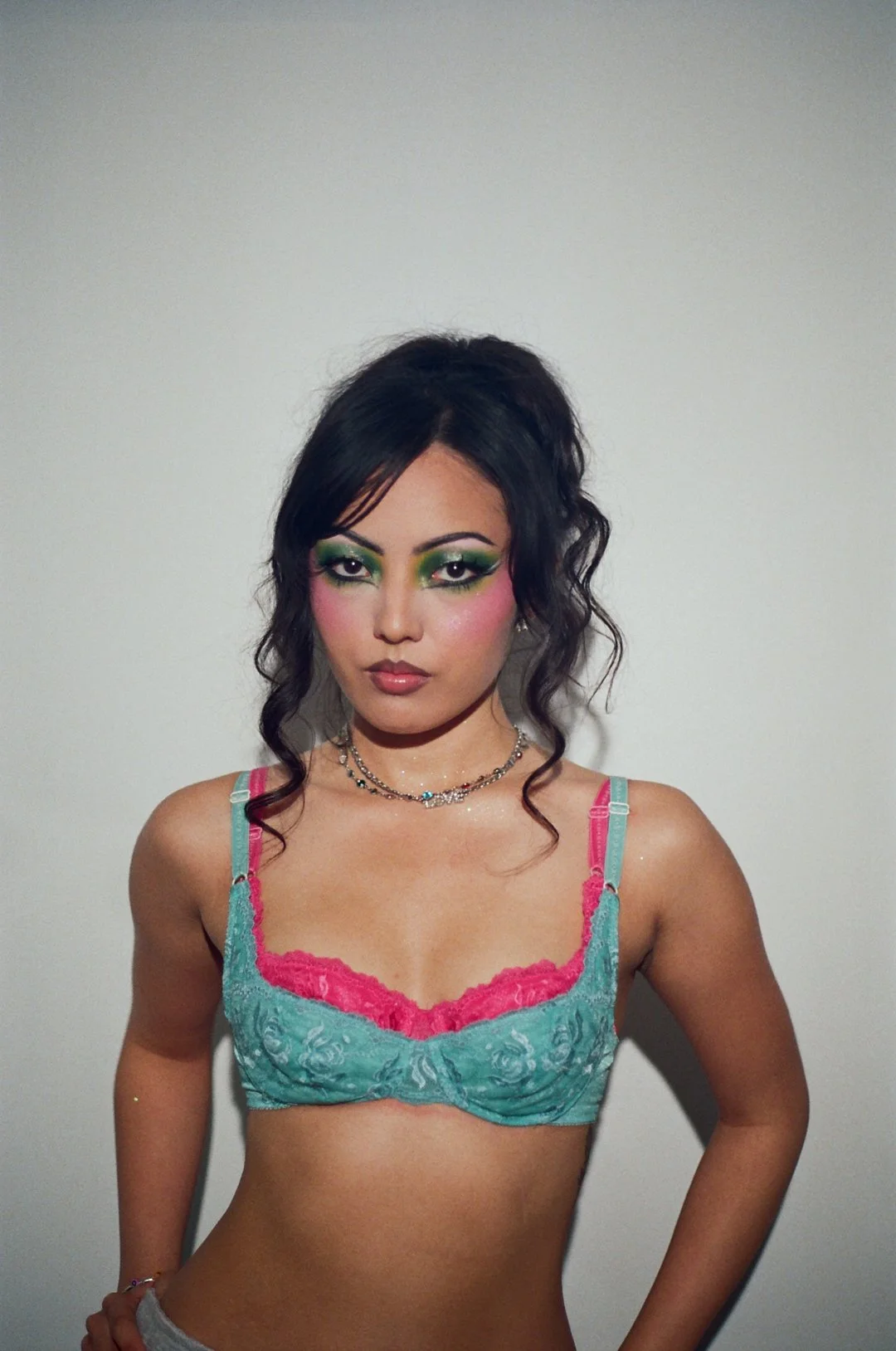A woman with dark hair and colorful makeup posing against a plain wall wearing a teal and pink lingerie top.