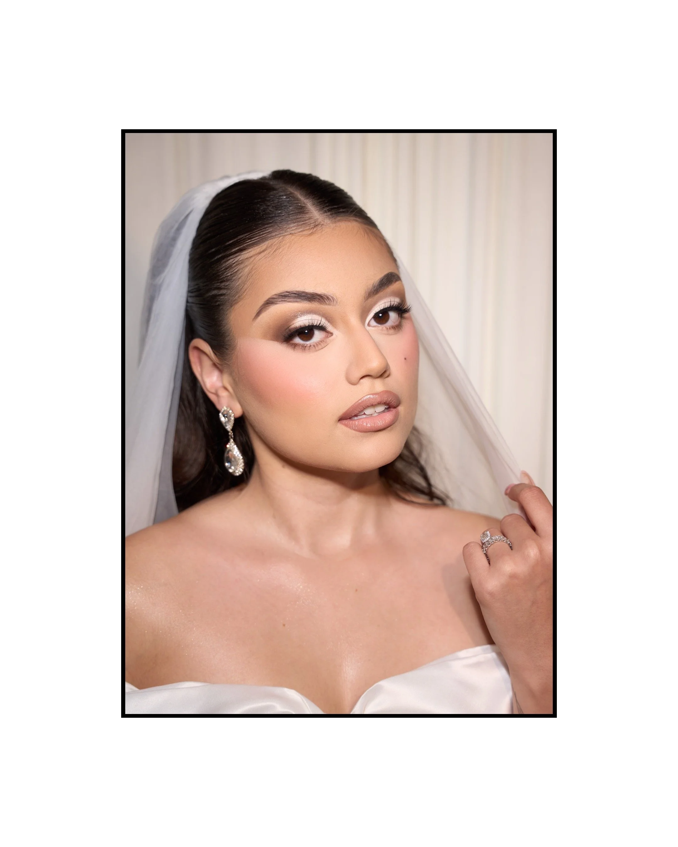 Close-up portrait of a young woman with makeup, earrings, and a veil, holding her hand near her face.