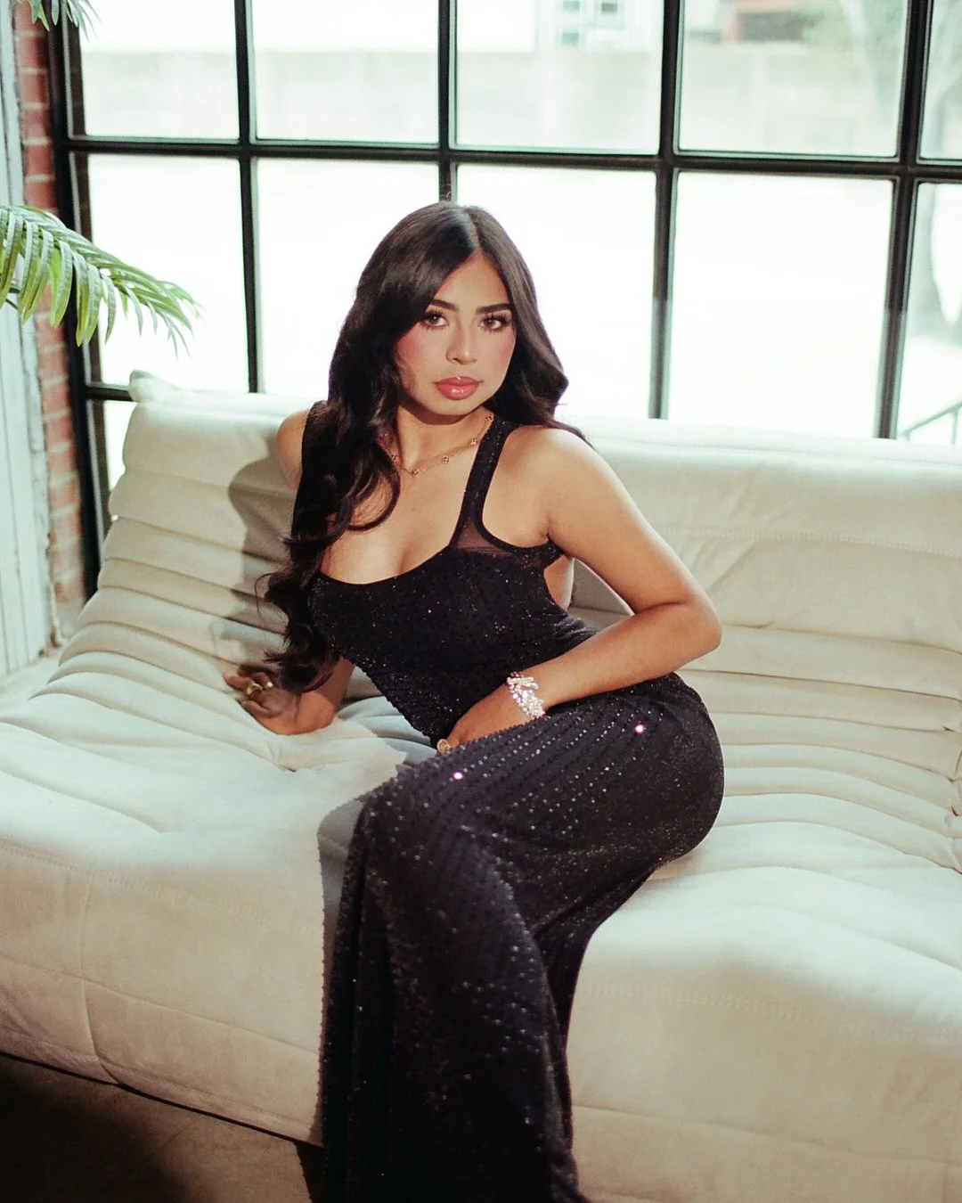 A woman in a black, sparkly dress sitting on a white couch in front of a large industrial-style window.