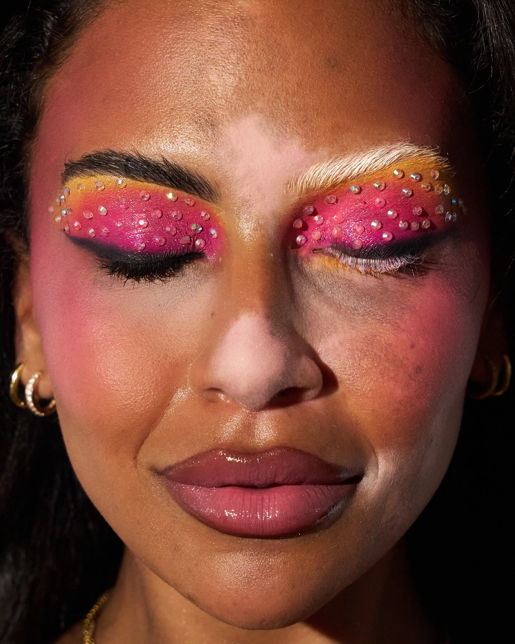 A woman with closed eyes wearing bright pink eyeshadow decorated with rhinestones and white false eyelashes, dark eyeliner, and soft pink lipstick. She has gold hoop earrings and a gold necklace.
