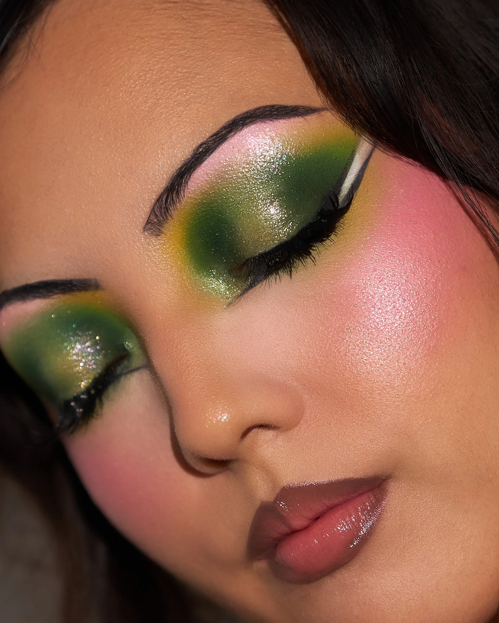 Close-up of a woman's face with vibrant, glittery makeup, featuring pink, yellow, and green eyeshadow, shimmery cheeks, and glossy lips.