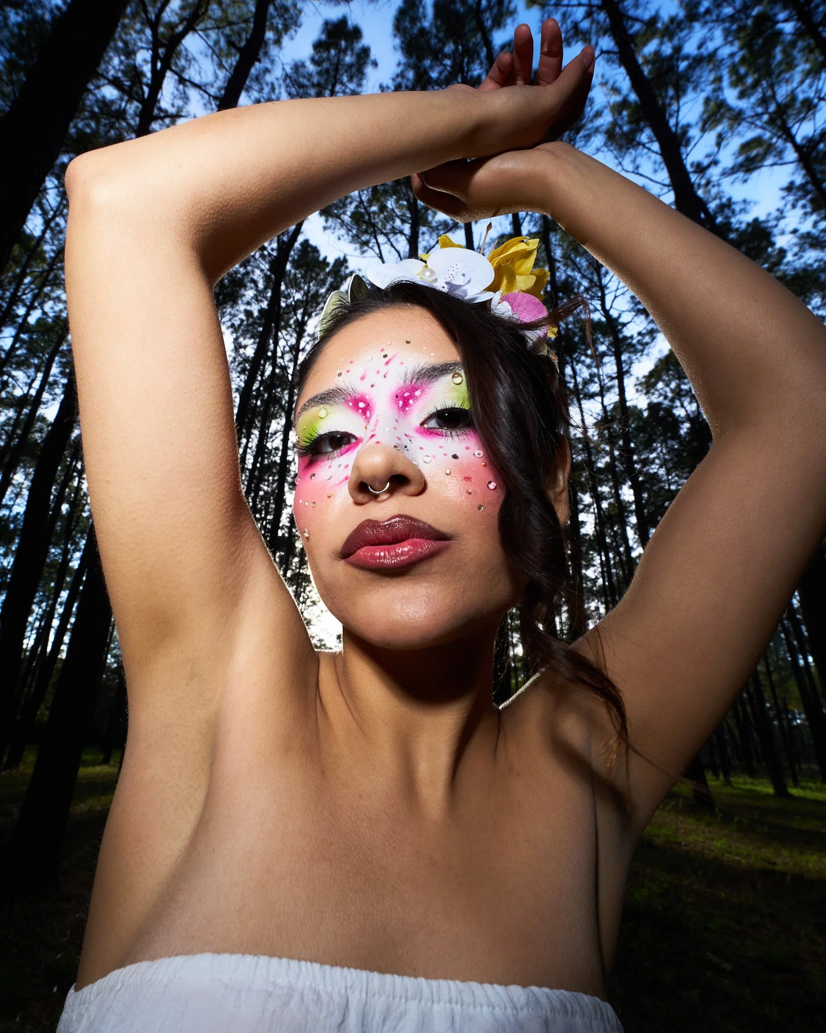 A woman with colorful makeup and rhinestones on her face, wearing a flower crown, poses with arms raised in a forest setting during twilight.