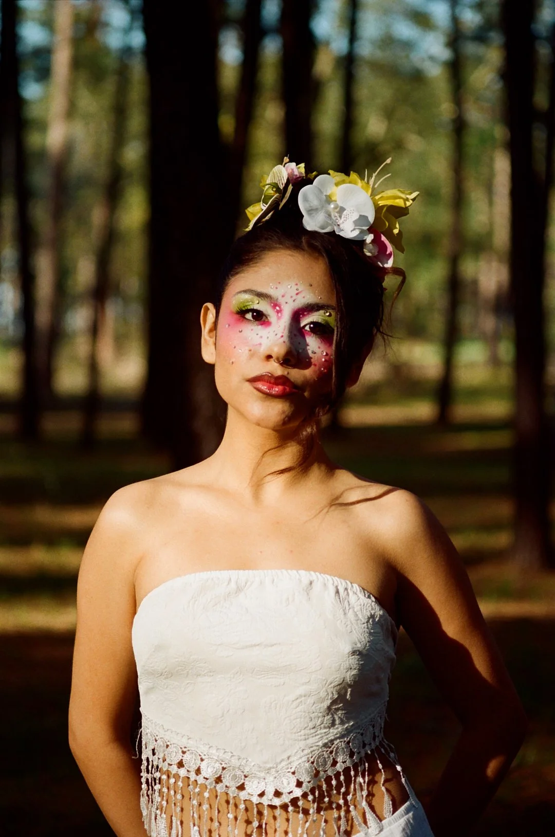 A woman with elaborate makeup and floral headpiece standing outdoors in a wooded area.