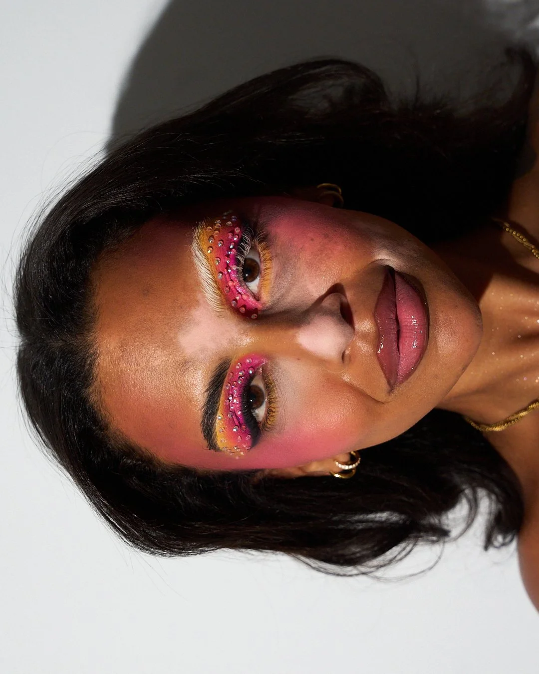 A woman with bold makeup featuring bright pink, yellow, and orange eyeshadow, rhinestones around her eyes, and glossy pink lips.