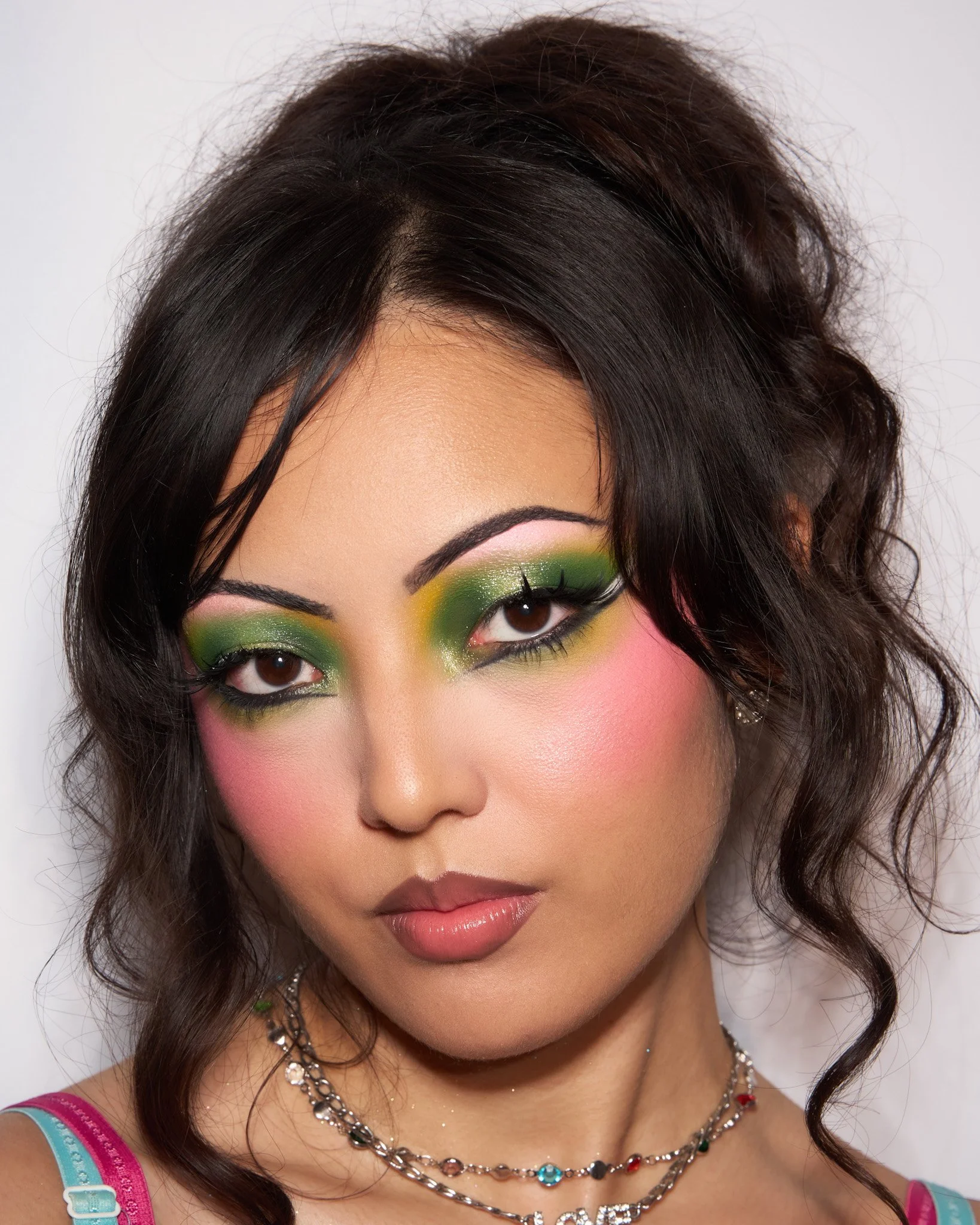 Close-up of a woman with dark, wavy hair wearing colorful makeup including green, pink, and yellow eyeshadow, and jewelry.