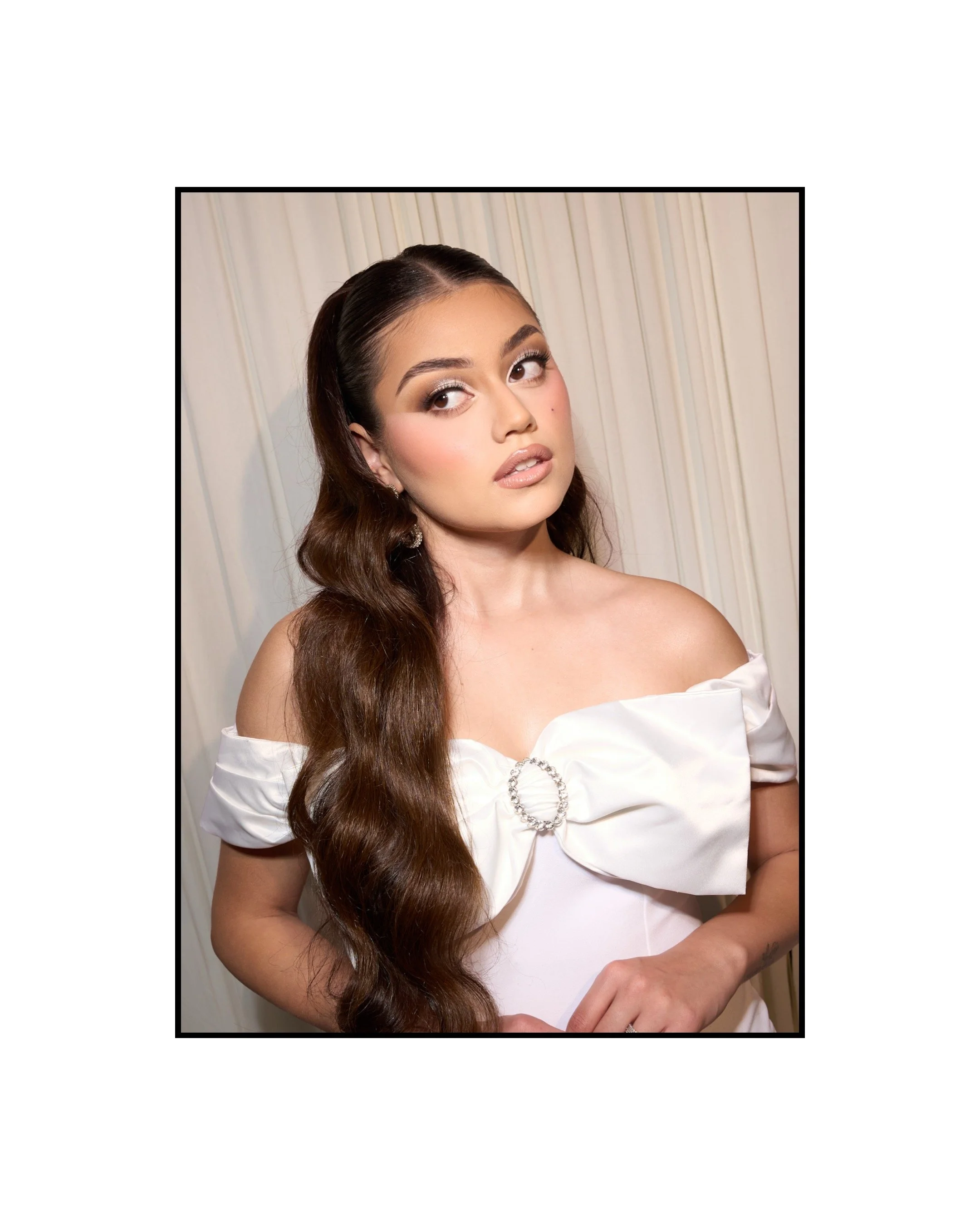 A young woman with long, wavy brown hair, wearing a white off-shoulder dress with a bow detail and a rhinestone ring, posing against a neutral curtain background.
