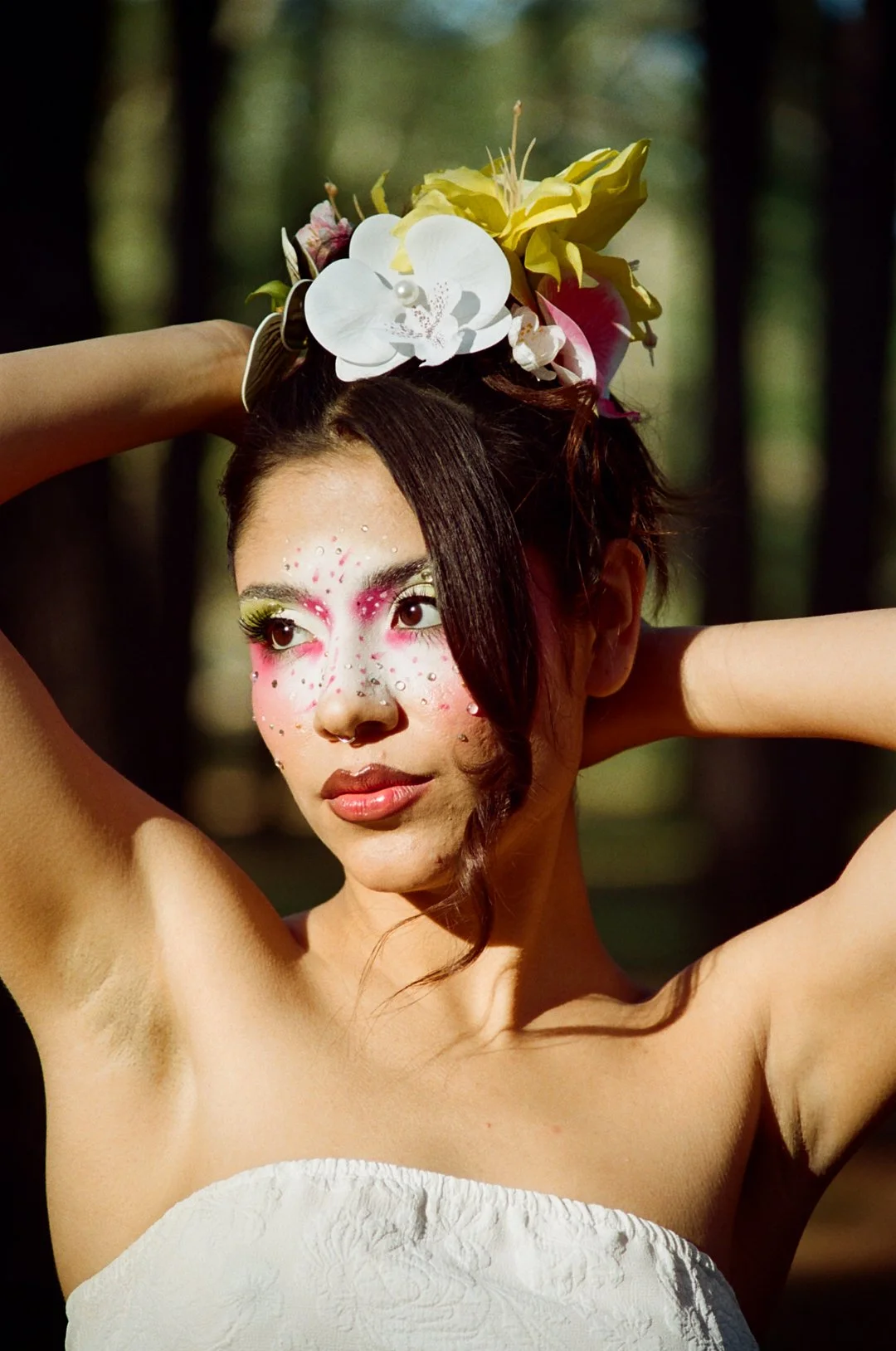 A woman with floral headpiece, colorful makeup with rhinestones, and a strapless white dress in a wooded outdoor setting.