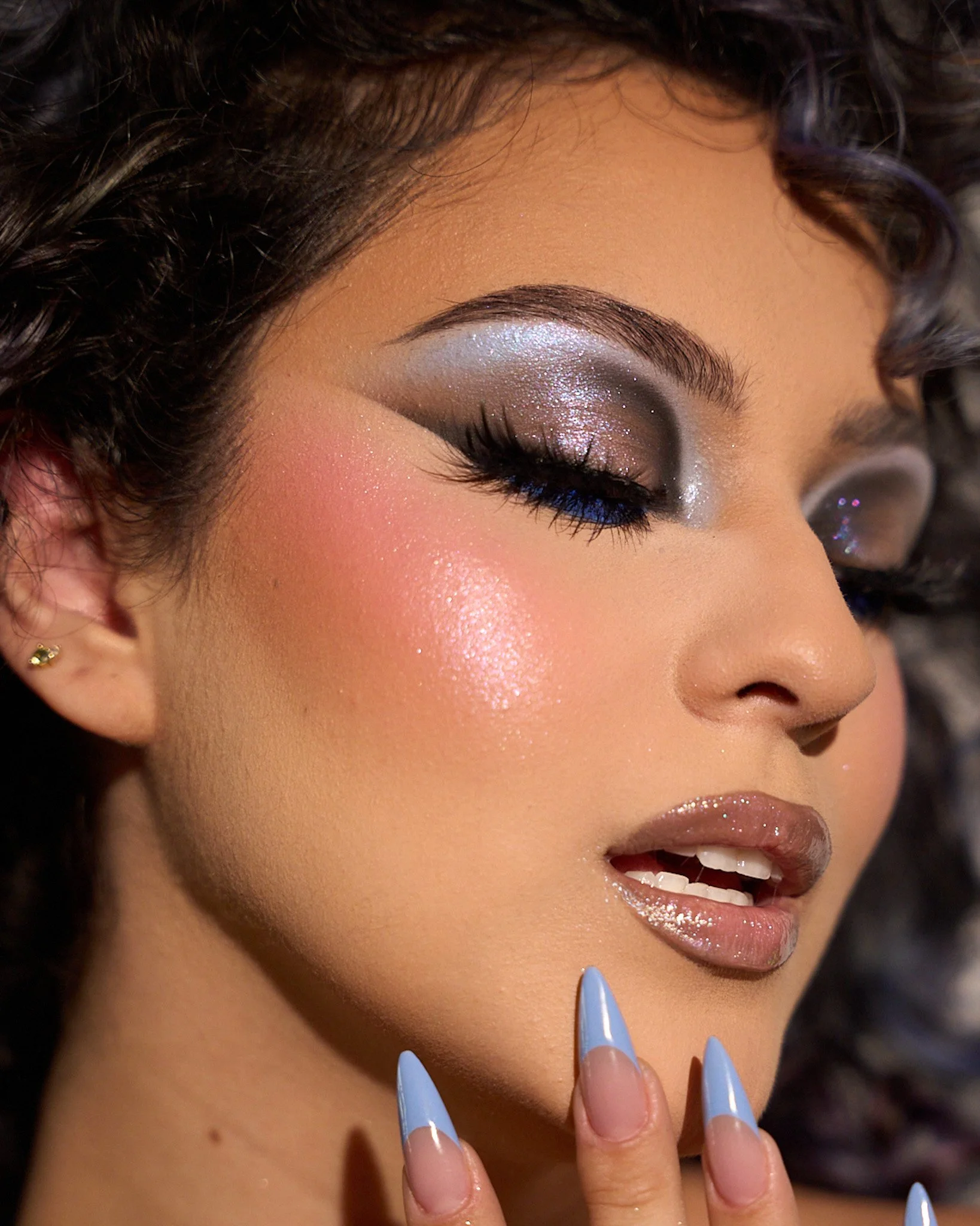 Close-up of a woman with glamorous makeup, including silver and black eyeshadow, blue eyeliner, and glossy lips, with her hand near her chin and nails painted light blue.