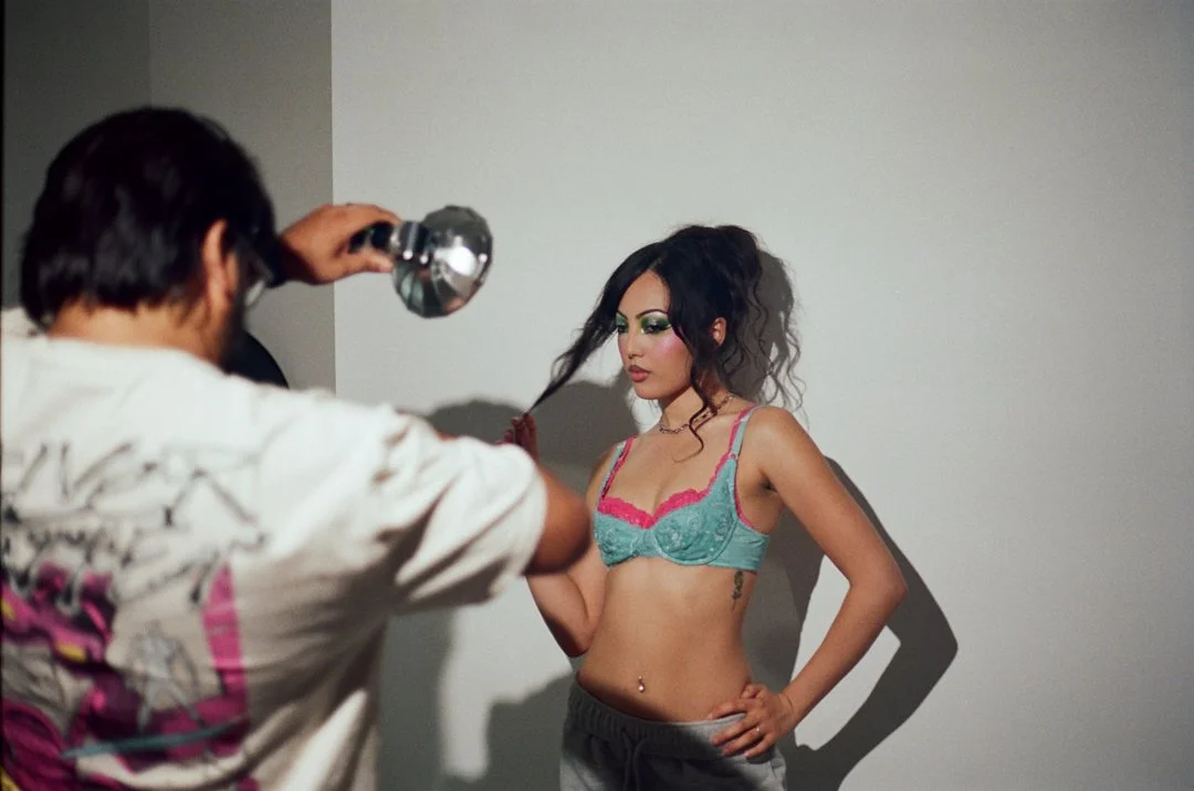 A woman with dark, curly hair in a messy ponytail wearing a colorful bra and gray pants is posing against a plain wall while a photographer captures her photo.