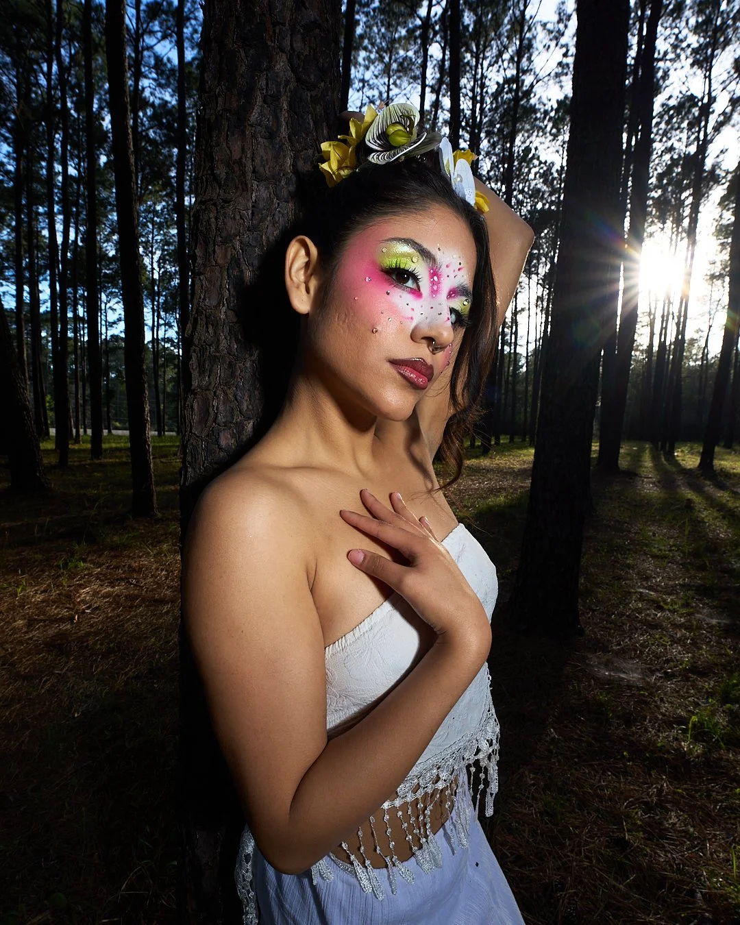 A woman with artistic makeup and floral headpiece posing in a forest at sunset.
