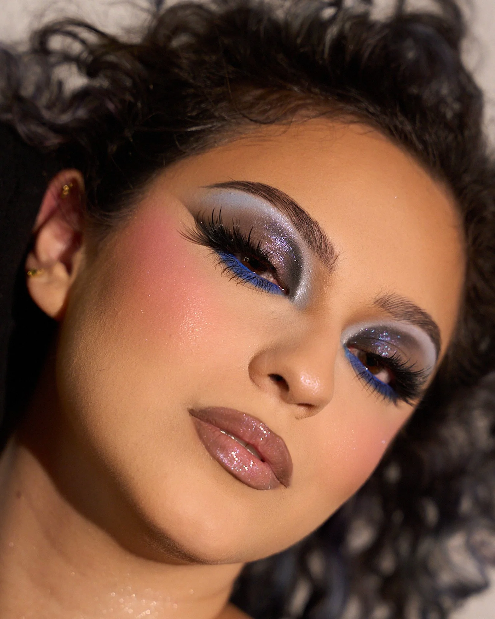 Close-up of a woman with glamorous makeup featuring shimmering silver eyeshadow, bright blue eyeliner, long lashes, and glossy lips. She has curly hair and multiple earrings.