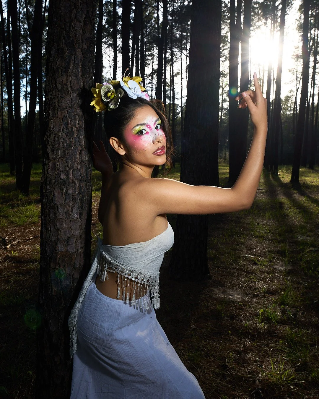 A woman with colorful makeup and floral headpiece standing in a forest, with sunlight streaming through trees.
