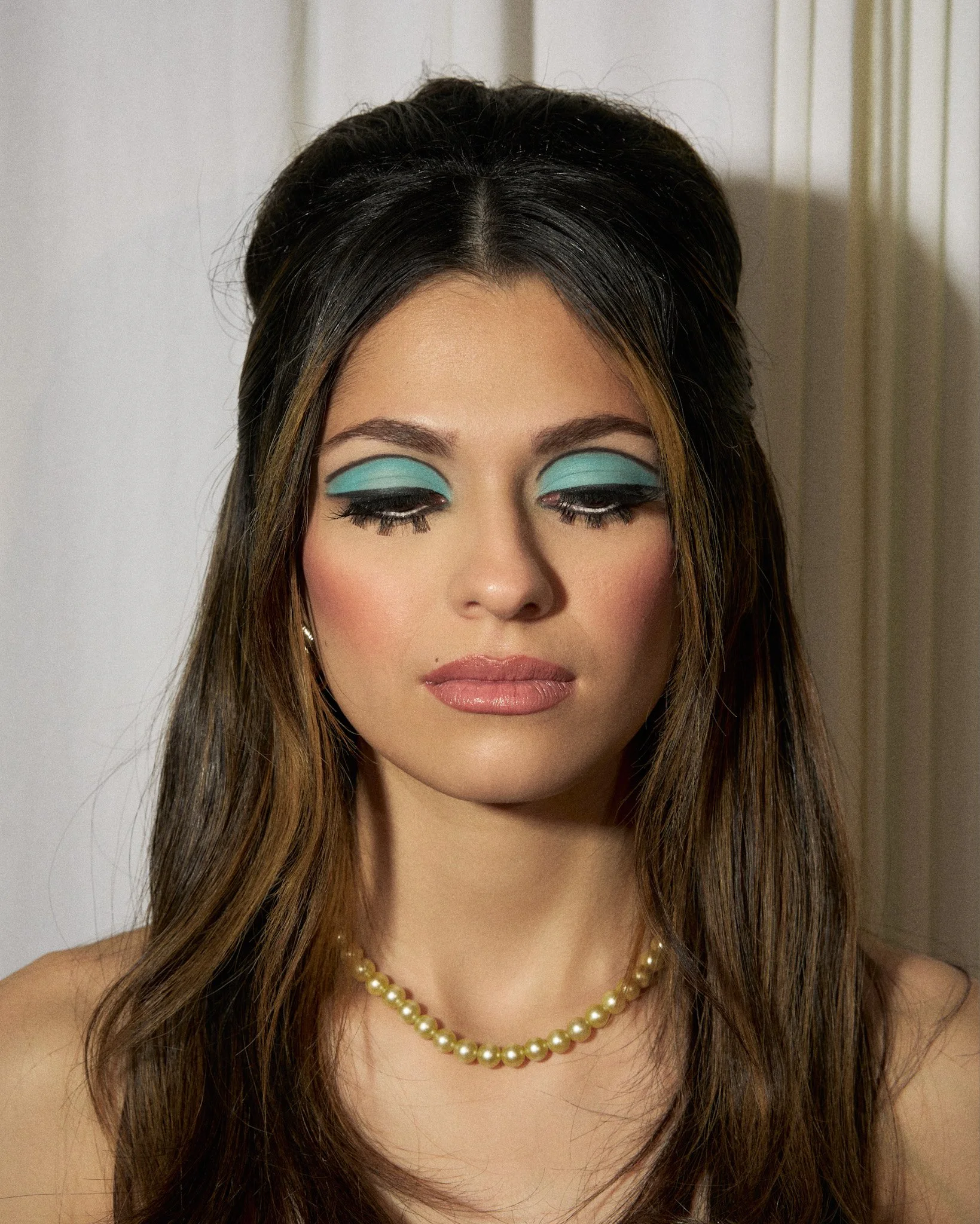 Close-up of a woman with makeup and a pearl necklace, with her eyes closed.