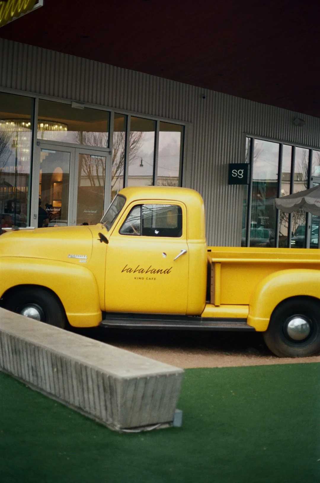 Yellow vintage truck parked outside a building with large glass windows, with the words "lalaland KIND CAFE" on the door, and a small sign with "SQ" on it.