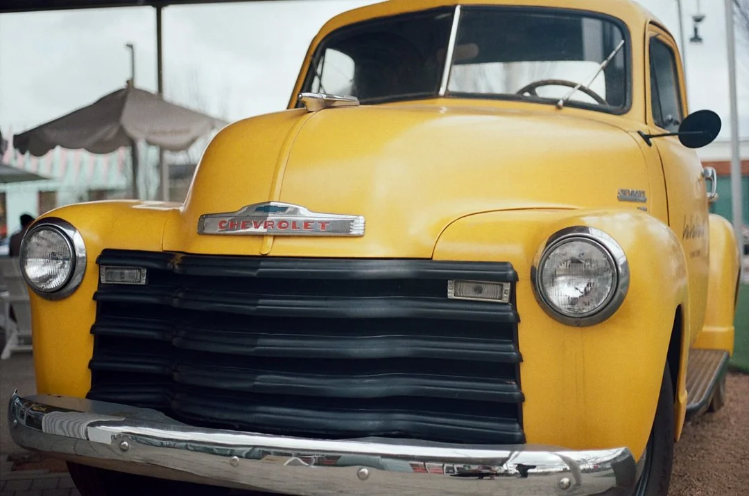 Yellow vintage Chevrolet truck parked outdoors with umbrellas in the background.