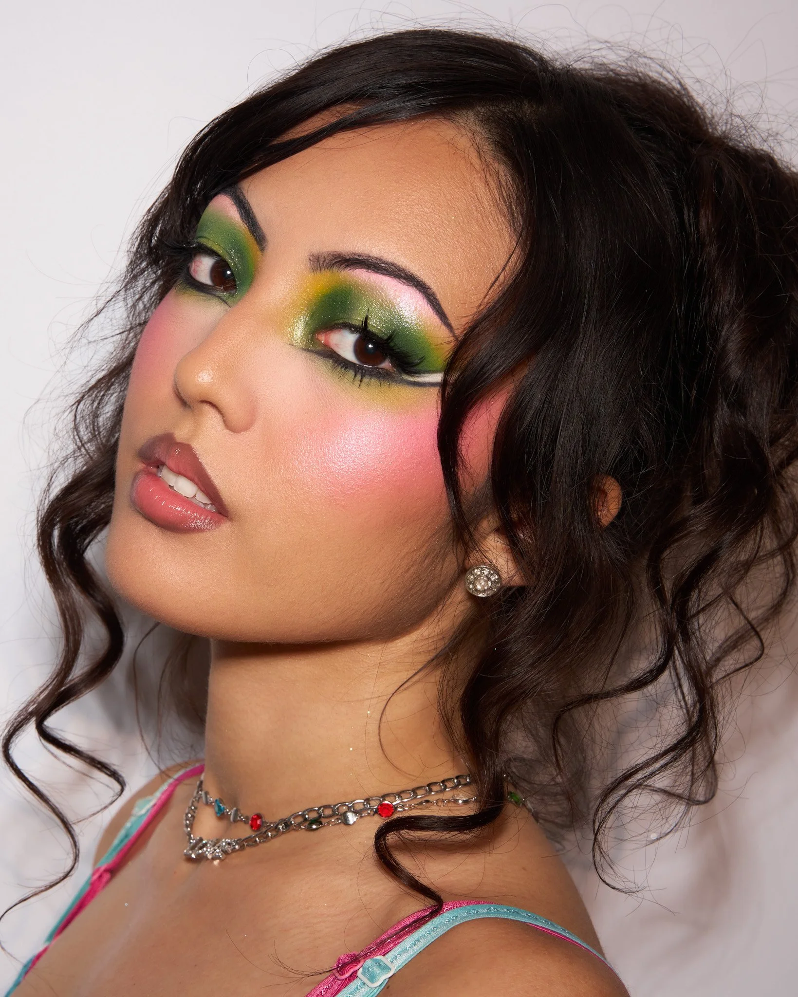 Close-up of a young woman with vibrant green and pink makeup, dark curly hair, diamond earrings, layered necklaces, and wearing a colorful top.