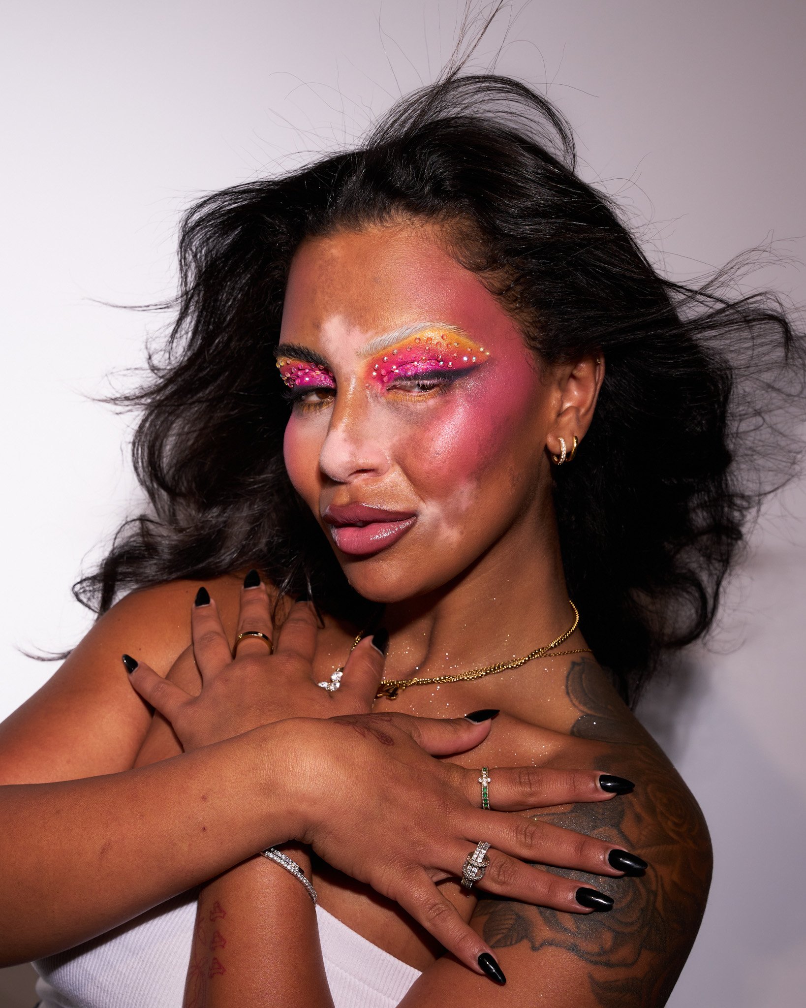 A woman with colorful makeup, including pink, orange, and yellow eyeshadow with rhinestones, and pink blush, posing with hands crossed over her chest, wearing jewelry and black nail polish.