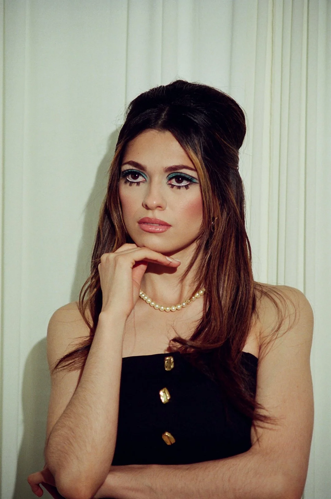 A young woman with long brown hair styled half-up, wearing bold makeup with blue eyeshadow and false eyelashes, a pearl necklace, and a black dress with gold buttons. She is standing against a white paneled wall, resting her chin on her hand, looking thoughtfully at the camera.