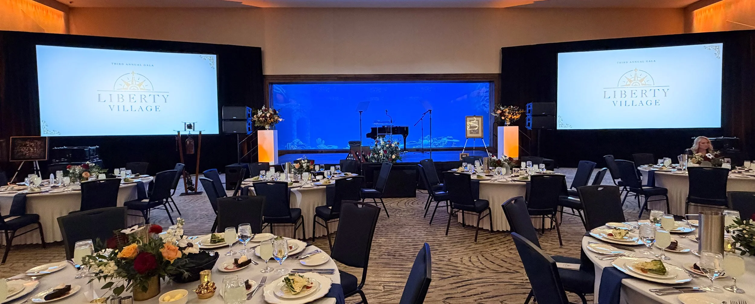 Elegant gala event with dual projector screens, piano on stage, floral centerpieces, and catered dining tables