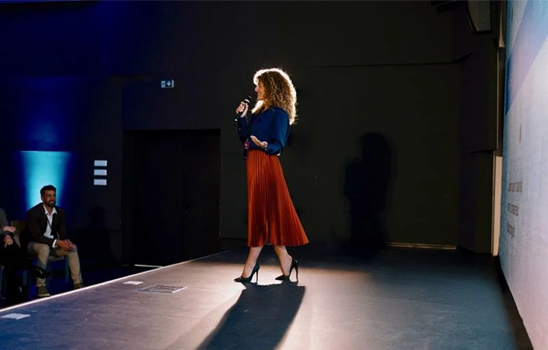 Woman presenter speaking on stage at a corporate event with LED wall behind her