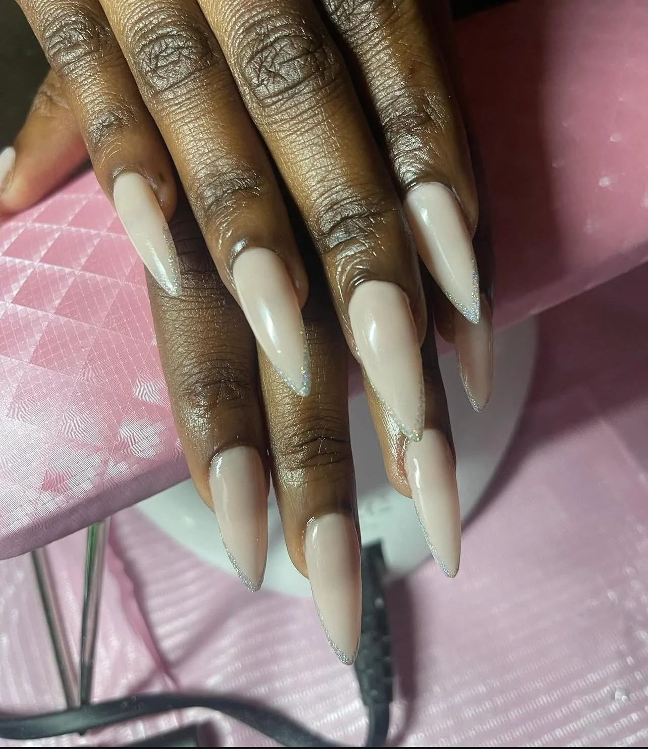 Close-up of long, sharp, cream-colored painted nails with a subtle sparkle, resting on a pink textured surface.