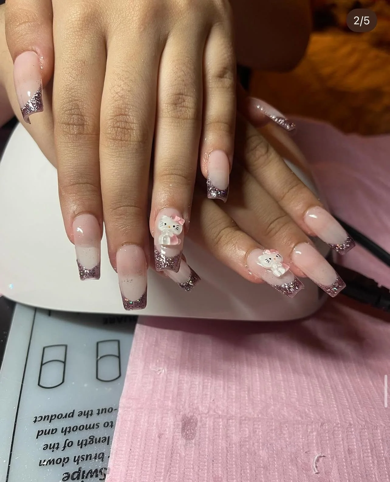 Close-up of manicured nails with pink and white gradient design, glitter accents, and 3D Hello Kitty nail art on ring fingers, with a pink paper towel underneath.