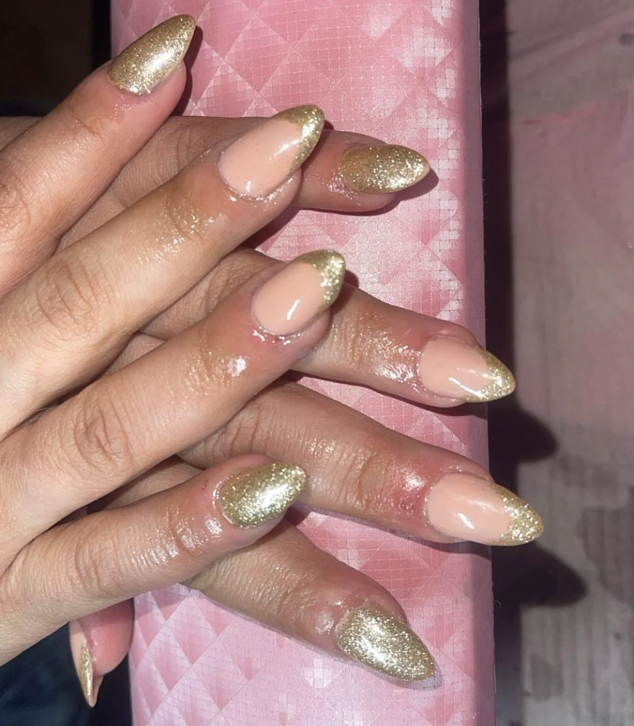 Close-up of a person's hand with gold glitter french-tip styled nails resting on a pink surface.