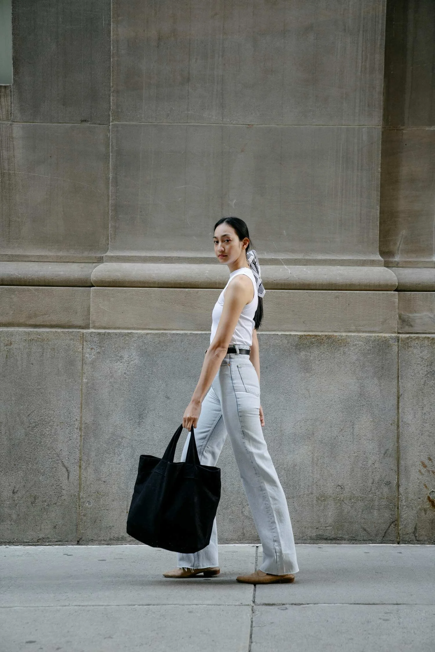 A young woman with dark hair tied back, wearing a white sleeveless top and light blue jeans, walking along a sidewalk in front of a large stone wall. She carries a black tote bag in her right hand and wears beige shoes.