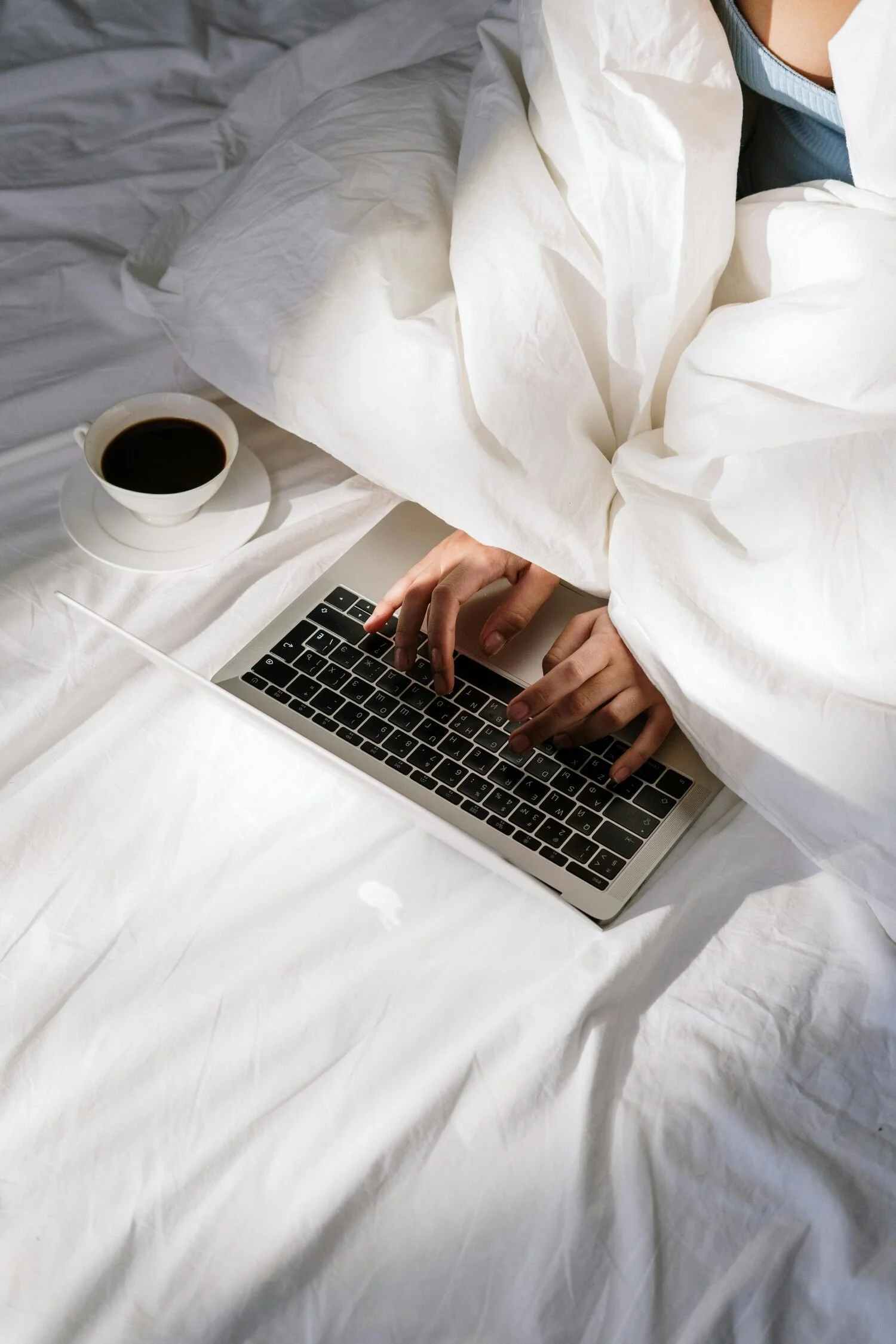 Person using a laptop on a bed with a cup of coffee nearby.