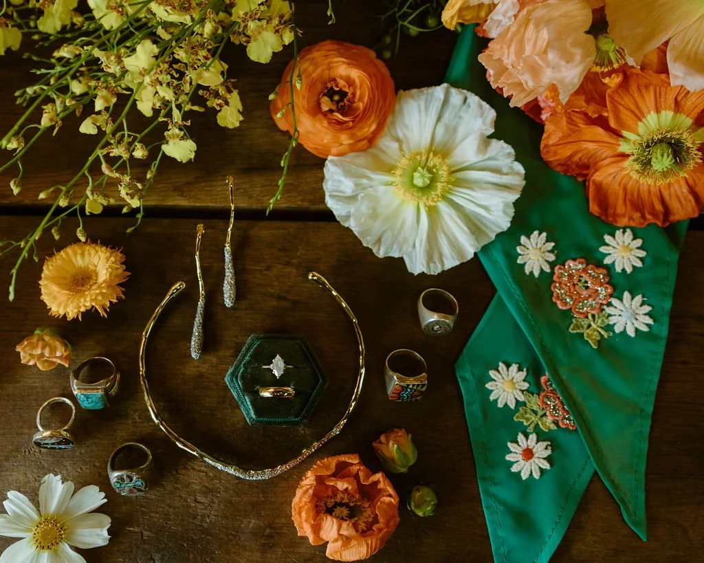 A collection of jewelry including rings, earrings, a necklace, and a ring box, surrounded by orange, white, and yellow flowers on a wooden surface with a green embroidered cloth.