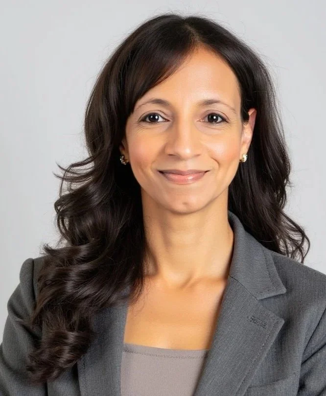 Professional woman with long, wavy dark hair, smiling, wearing a gray blazer and earrings, against a plain light background.