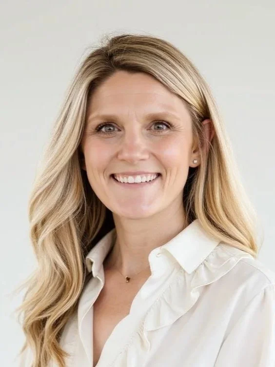 A woman with long blond hair smiling, wearing a white blouse and small earrings, standing against a plain light background.