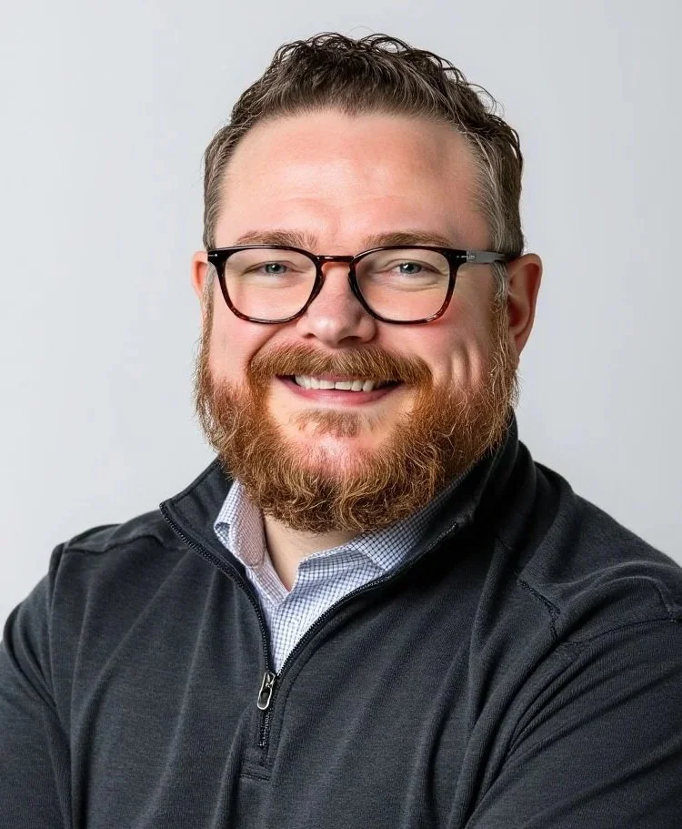 A smiling man with glasses, a beard, and short hair wearing a black zip-up jacket over a collared shirt posing against a plain background.