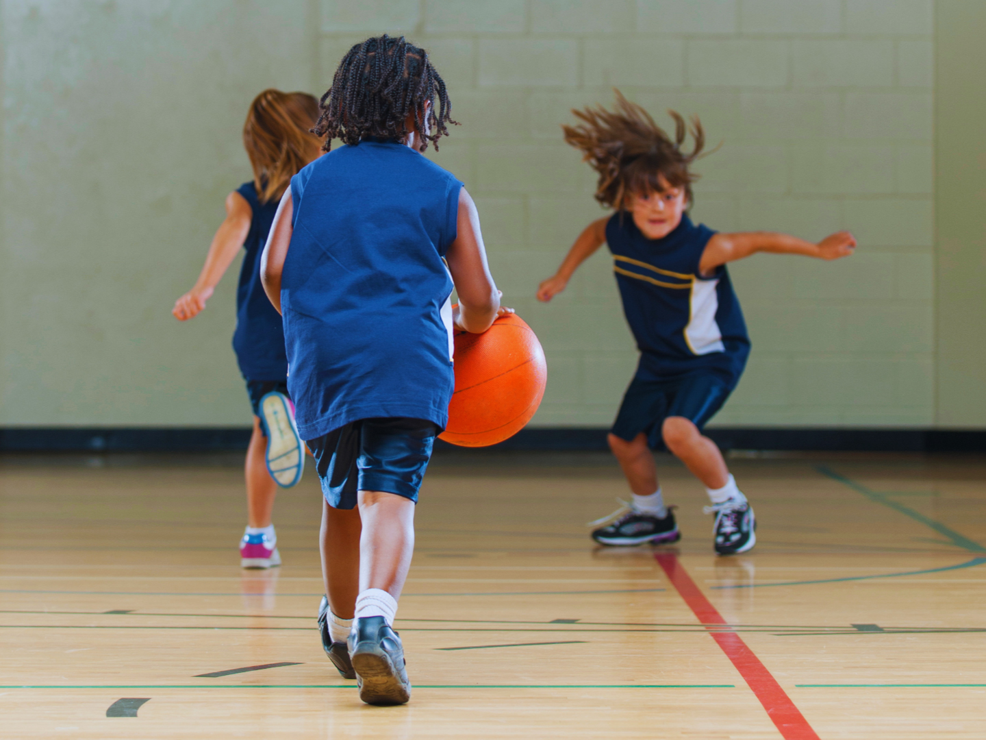Kids playing basketball in a gymnasium, with one child dribbling the ball and another preparing to defend.