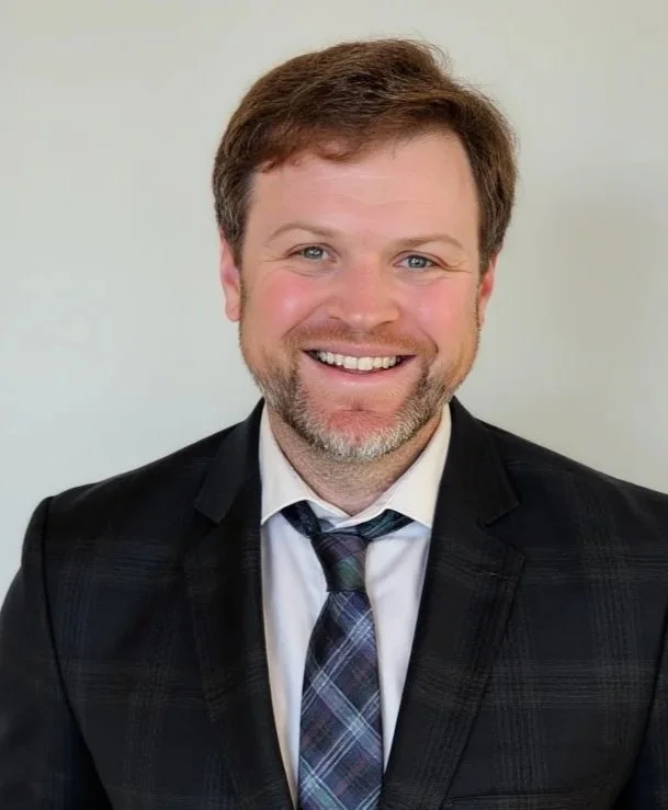A smiling man with light brown hair, a beard, wearing a dark suit, white shirt, and plaid tie, standing against a light background.
