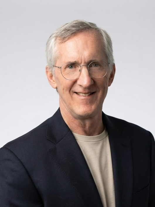 A man with gray hair and glasses wearing a dark blazer and a light-colored shirt, smiling at the camera against a plain white background.