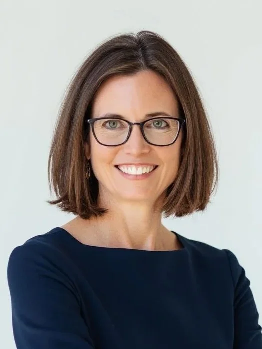 A smiling woman with shoulder-length brown hair, wearing glasses and a navy blue top, standing against a light background.