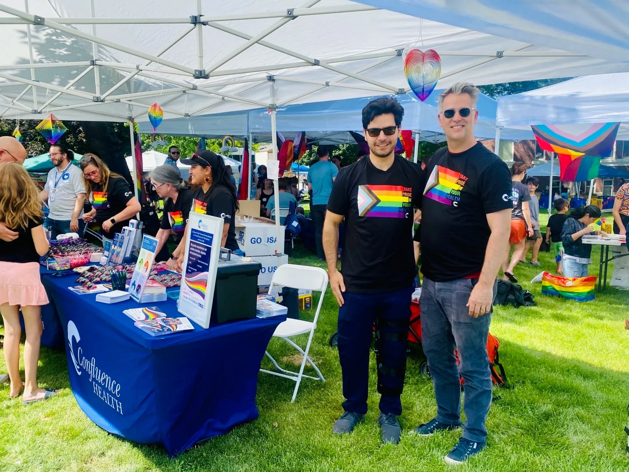 Two men wearing black Pride T-shirts standing together at an outdoor pride event, with a booth decorated with rainbow-themed items and other people in the background.