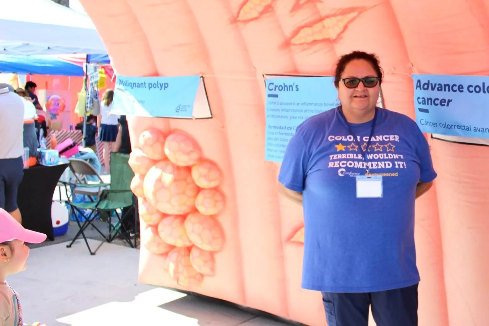 A woman wearing sunglasses and a blue t-shirt with a colon cancer awareness message, standing in front of an educational display about colon cancer at an outdoor event.