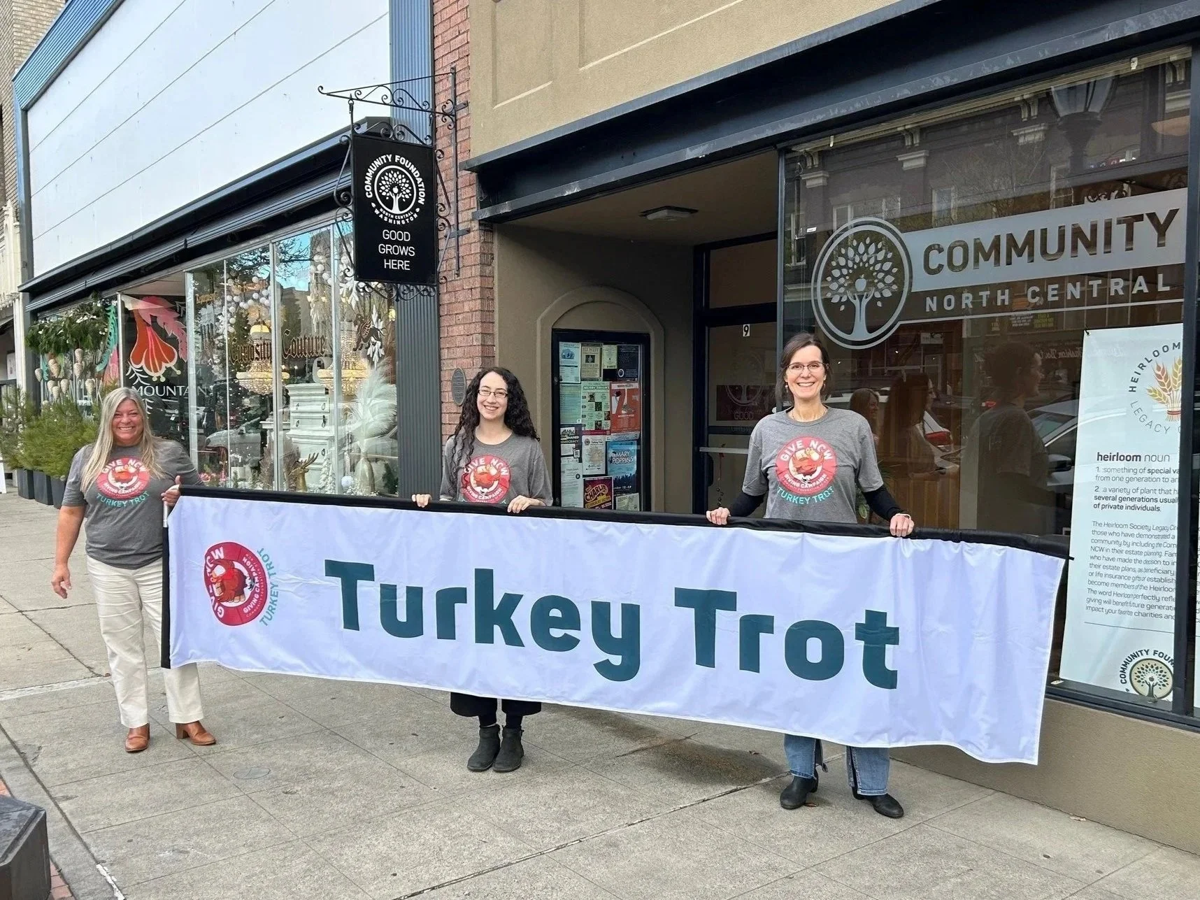 Three women standing on the sidewalk holding a large banner that reads 'Turkey Trot' in front of a storefront with a sign that says 'Community North Central'.