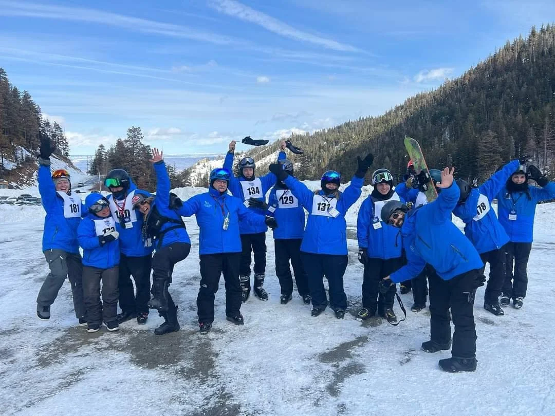 A group of people dressed in blue jackets and helmets, celebrating on a snowy landscape with mountains and trees in the background, some holding skis or snowboards.