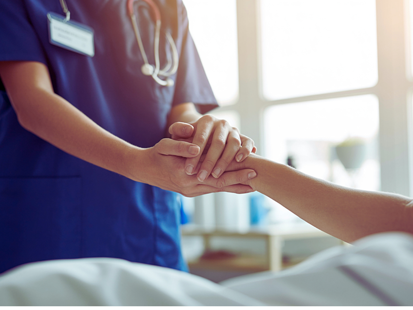 A healthcare professional, wearing blue scrubs and a stethoscope, gently holds a patient's hand in a bright clinical setting.