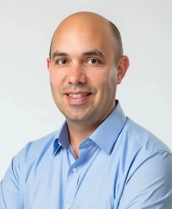 Portrait of a bald man in a light blue button-up shirt smiling at the camera against a plain white background.