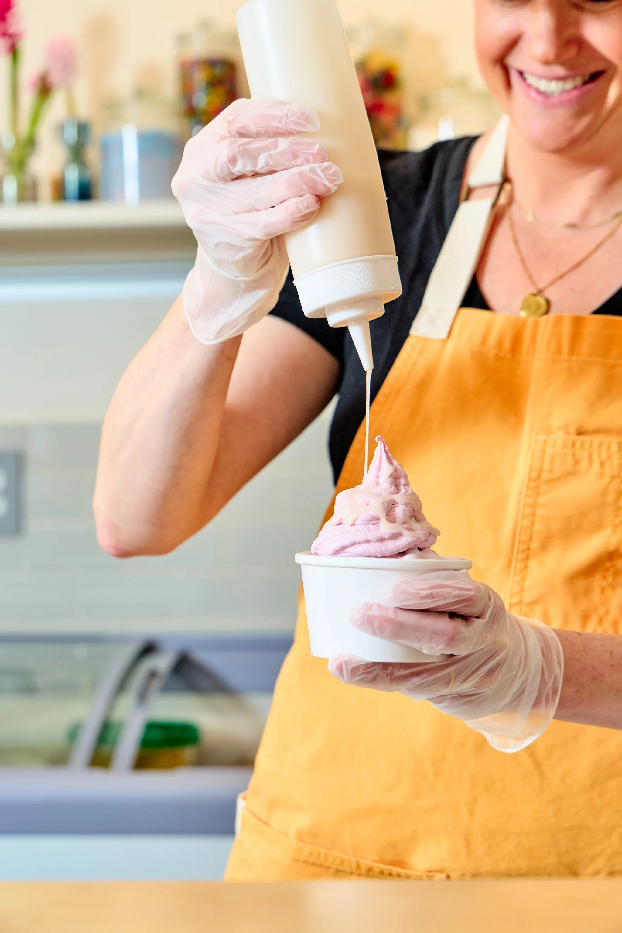 A woman in an orange apron and gloves is drizzling housemade syrup onto pink soft serve ice cream from a bottle onto a cup.