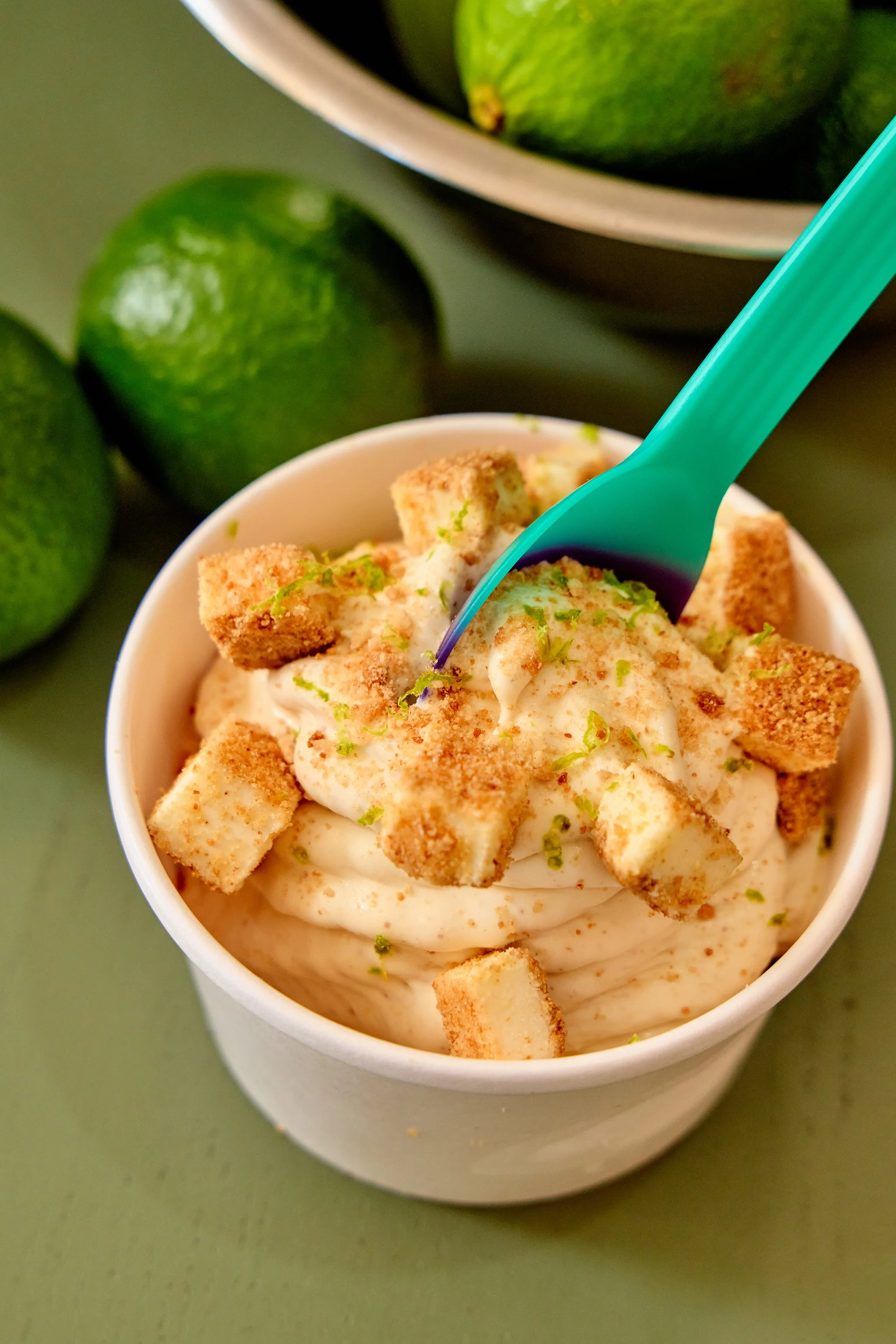 Cup of creamy frozen dessert topped with cheesecake bites and lime zest, with whole limes in the background.
