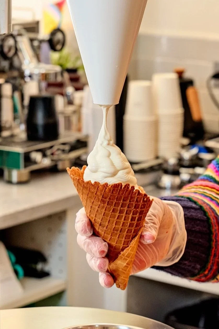 A hand wearing a colorful knitted sweater and gloves is holding a waffle cone topped with soft serve vanilla ice cream, with a cream dispensing from a cone-shaped container above.