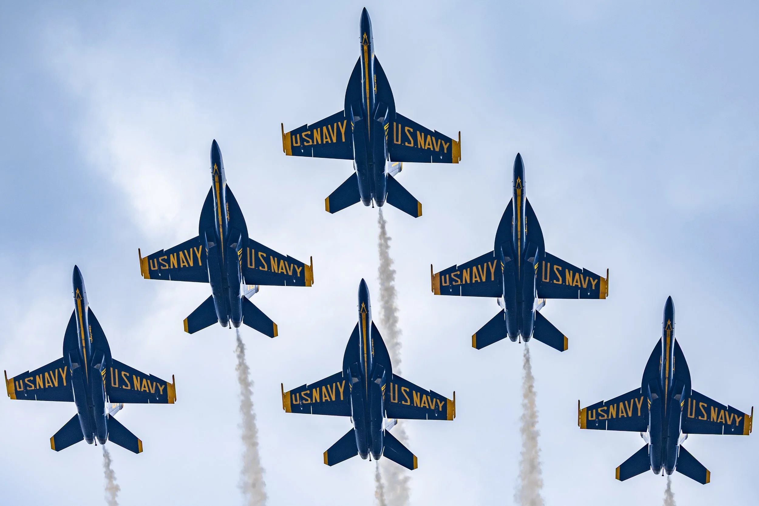 Multiple Blue U.S. Navy fighter jets flying in formation with smoke trails against a cloudy sky.