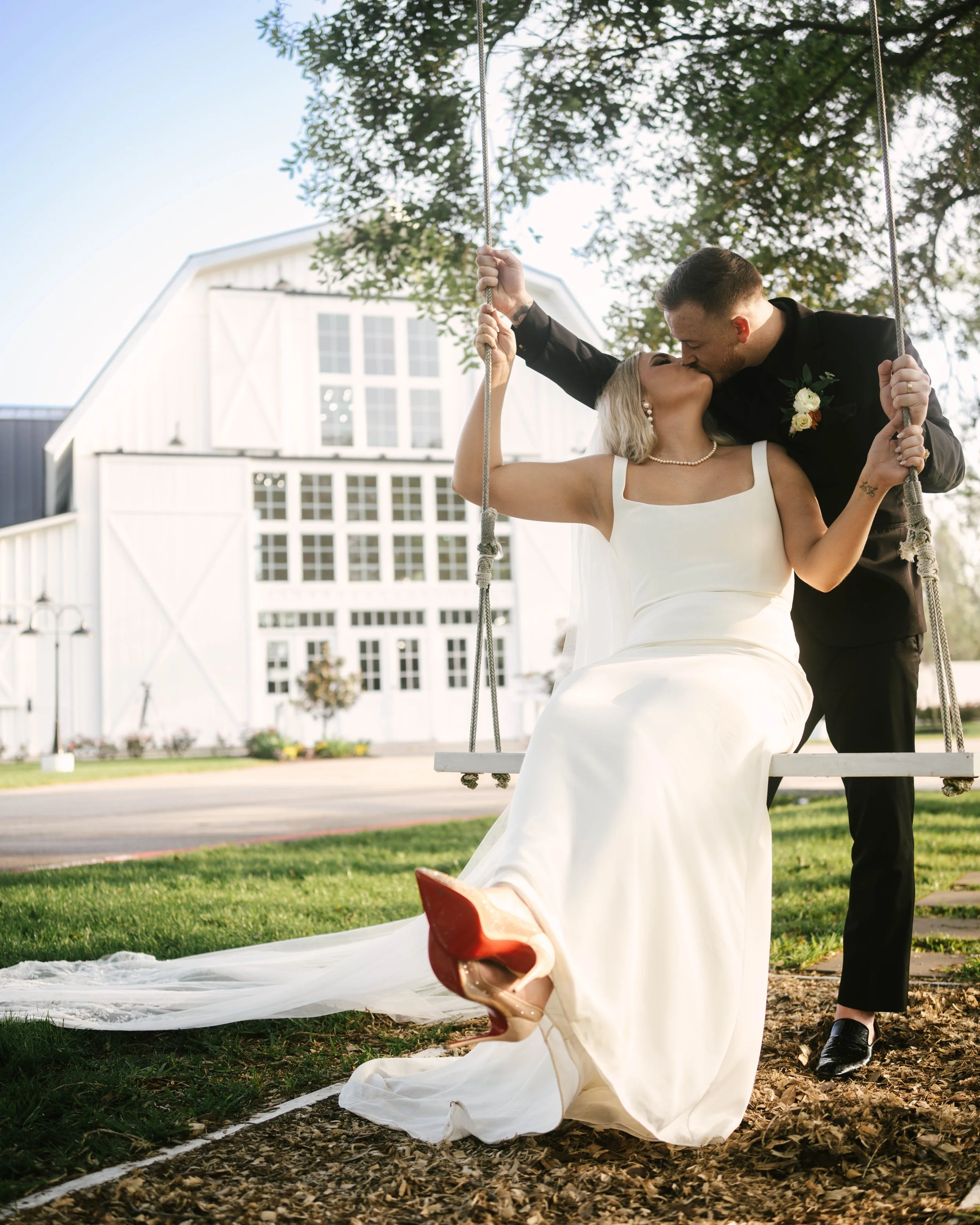A bride and groom share a kiss on a swing in front of a white barn, with the bride wearing a white wedding dress and red shoes, and the groom in a black suit.